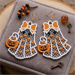 May include: A pair of white lace ghost earrings featuring orange embroidered bows and pumpkin buckets. Each ghost has a large orange bow at the top and small orange accents throughout the lace pattern. The earrings hang from gold-toned hooks.
