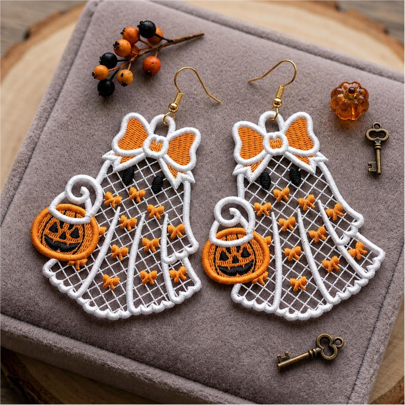May include: A pair of white lace ghost earrings featuring orange embroidered bows and pumpkin buckets. Each ghost has a large orange bow at the top and small orange accents throughout the lace pattern. The earrings hang from gold-toned hooks.