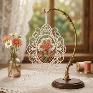 Puede incluir: Un adorno de encaje de color crema con forma ovalada que presenta un diseño floral bordado de margaritas blancas y coral. La pieza de encaje cuelga de un soporte de metal en tono latón antiguo con una base de madera circular y volutas.