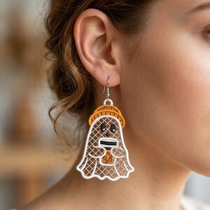 May include: A dangle earring featuring a white ghost character with a brown grid pattern, wearing an orange beanie and holding a brown bubble tea cup with black boba pearls. The ghost has black oval eyes and a small mouth, suspended from a silver hook.