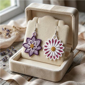 Pode incluir: Um par de brincos pendentes assimétricos com desenhos florais bordados. Um brinco é uma flor roxa de pétalas múltiplas com centro amarelo, enquanto o outro é uma flor branca e magenta tipo margarida com centro amarelo, ambos em ganchos prateados.