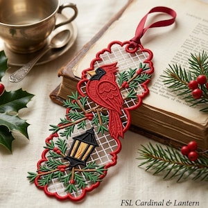 May include: A red cardinal bird and black lantern embroidered ornament with a white grid background, green pine branches, and red berries. The scalloped-edge design features a red ribbon loop at the top and the text FSL Cardinal & Lantern.