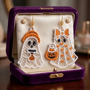 Puede incluir: Un par de pendientes bordados con forma de fantasma blanco y detalles en naranja, uno sosteniendo una bebida boba y el otro una calabaza, presentados en un joyero de terciopelo morado con dos pequeños pendientes de perlas sobre fondo crema.