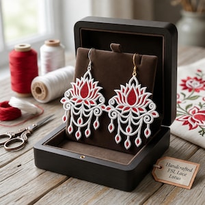 May include: A pair of white and red lotus-shaped lace earrings displayed on a brown velvet stand inside a dark wooden box. The earrings feature intricate scrollwork and dangling elements. A tag attached to the box reads Handcrafted FSL Lace Lotus.