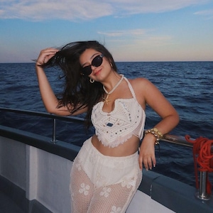 Op de afbeelding: Een witte gehaakte haltertop en bijpassende broek met bloemenpatroon. De persoon draagt een zonnebril en sieraden, leunend tegen een bootreling met de oceaan op de achtergrond.