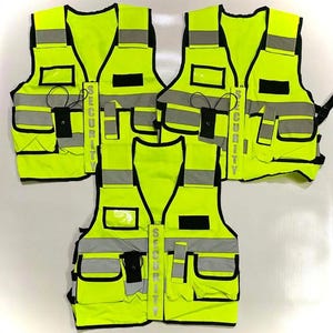 Op de afbeelding: Verschillende goed zichtbare veiligheidsvesten in felgeelgroen. Elk vest heeft reflecterende zilveren strepen, meerdere zakken en het woord "SECURITY" verticaal gedrukt. Ze hebben zwarte biezen en een klein zakje voor een radio of ander apparaat.