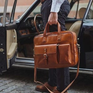May include: A tan leather briefcase featuring two top handles, a detachable shoulder strap, and a front pocket secured with a buckle strap. The bag has a rectangular shape with visible stitching, metal hardware, and a smooth, polished finish.
