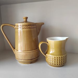 Op de afbeelding: Een vintage keramische koffiepot en creamer set in een warme mosterdgele kleur. De koffiepot heeft een deksel en een lange handgreep, met een decoratieve band rond de boven- en onderkant. De creamer heeft een ruitpatroon aan de onderkant.