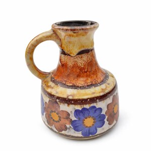 Op de afbeelding: Een keramische kan met een handvat, met een bloemenontwerp. De kan heeft een bruin, oranje en crèmekleurig kleurenschema, met blauwe en bruine bloemen rond de basis. De bovenkant en het handvat zijn lichtbeige.