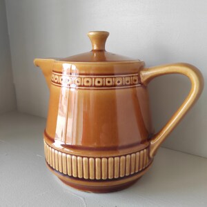 May include: A brown ceramic jug with a lid and handle. The jug has a glossy finish and is decorated with a geometric pattern around the top and bottom. The handle is curved, and the spout is small.