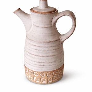 Op de afbeelding: Een handgemaakte keramische kan met een deksel. De gebroken witte kan heeft een cilindrisch lichaam met horizontale ribbels en een decoratieve band van cirkels en vormen aan de basis. Het heeft een gebogen handvat en een tuit.