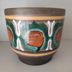 Op de afbeelding: Een keramische plantenpot met een bruine basis en rand, met abstracte oranje, groene en witte ontwerpen. De pot heeft een cilindrische vorm met een licht uitlopende basis en een decoratieve band van gele en bruine strepen.