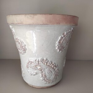 Op de afbeelding: Een taps toelopende keramische plantenpot met een glanzende witte craquelé glazuurafwerking. De buitenkant is versierd met verhoogde, getextureerde paisley-motieven in een roodbruine tint. De brede, platte rand is ongeglazuurd, wat de natuurlijke kleur van terracotta onthult.