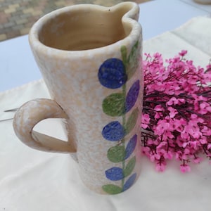 Op de afbeelding: Een beige keramische kan met een handvat, versierd met blauwe en groene bloempatronen. De kan heeft een gespikkelde textuur en een unieke, golvende rand. Een boeket roze bloemen staat op de achtergrond.