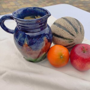 Pode incluir: Um jarro de cerâmica azul feito à mão com alça, bico e esmalte colorido. Ao lado, um melão cantalupo, uma laranja e uma maçã vermelha, dispostos sobre um pano branco. O jarro tem cerca de 18 cm de altura.
