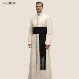 May include: A cream-colored robe with a mandarin collar and black buttons. The robe has black trim and is paired with a wide black sash with fringe. The image includes the text "CANDLEWICK TAILORING".