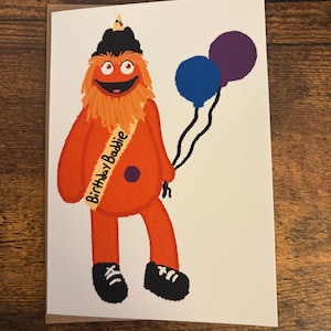 Op de afbeelding: Een wenskaart met een illustratie van een oranje, harig monster met een zwarte hoed, zwarte schoenen en een sjerp met de tekst Birthday Baddie. Het monster houdt een blauwe en een paarse ballon vast tegen een effen witte achtergrond.