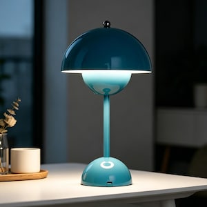 Puede incluir: Una lámpara de mesa de color turquesa con una pantalla y base redondeadas. La lámpara tiene un tallo delgado y una pequeña bola plateada en la parte superior. La lámpara está sobre una mesa blanca.