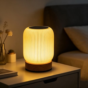 May include: A warm, illuminated table lamp with a cream-coloured, ribbed shade. The lamp is set on a wooden base and provides a soft glow. The lamp is approximately 15 cm tall and is placed on a white bedside table.