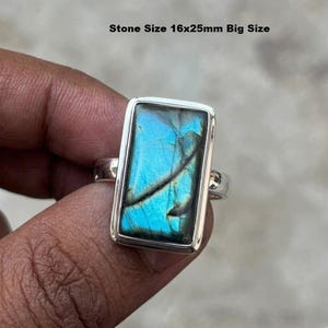 May include: A silver ring with a rectangular, iridescent blue gemstone. The stone displays a vibrant play of colour, with flashes of blue and green. The text "Stone Size 16x25mm Big Size" is visible above the ring.