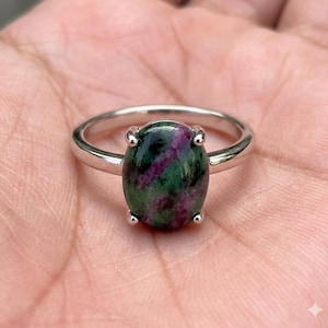 May include: A silver ring with an oval gemstone displaying green and purple colours. The gemstone is the central feature, set in a simple band. The ring is displayed against a skin-toned background.