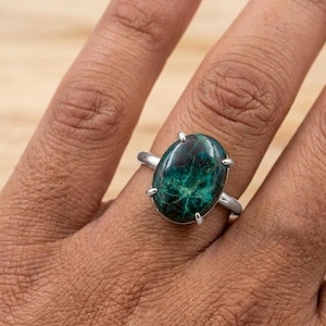 Puede incluir: Un anillo de plata con una piedra preciosa ovalada de color verde oscuro con intrincados patrones. La piedra preciosa está sujeta por cuatro garras. El anillo se muestra en el dedo de una persona.