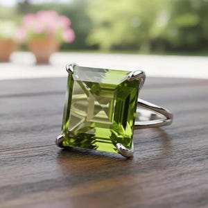 Puede incluir: Un anillo de plata con una gran piedra preciosa rectangular de color verde claro. La piedra preciosa está engastada en un engaste de cuatro garras, y la banda tiene un diseño dividido. El anillo se muestra sobre una superficie de madera.
