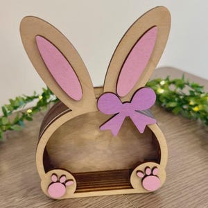 Può includere: Scaffale in legno a forma di coniglietto pasquale con dettagli rosa. Lo scaffale presenta orecchie di coniglio, un fiocco e impronte di zampe. Le orecchie e le impronte sono rosa e il fiocco è di una tonalità di viola più chiara.