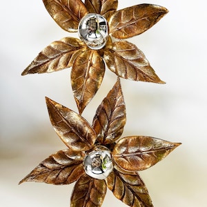Puede incluir: Dos apliques de pared decorativos con forma de flor. Cada aplique presenta un acabado dorado metálico con pétalos detallados en forma de hoja que rodean un orbe plateado reflectante. Diseñados para montaje en pared.