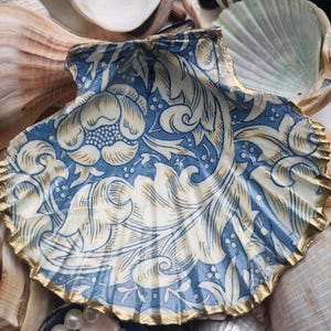 May include: A painted scallop shell featuring a blue and cream floral design, with a gold painted edge. The background includes various seashells, creating a coastal aesthetic.
