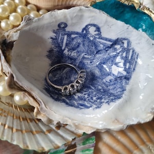 May include: A silver ring with five clear stones sits within a white seashell featuring a blue and white illustration. The seashell is surrounded by other shells and a string of faux pearls, creating a coastal theme.