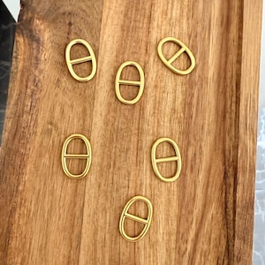 Peut inclure: Cinq boucles en métal doré, chacune composée d'un cadre ovale avec une barre centrale horizontale. Ces petits composants de quincaillerie lisses sont disposés de manière éparse, présentant une finition polie et un design uniforme.