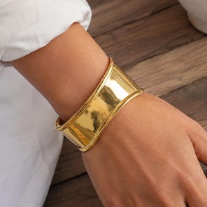 Peut inclure: Un large bracelet manchette doré et poli avec une surface lisse et réfléchissante aux bords légèrement incurvés. Le bracelet présente un design en bande solide et continue qui entoure le poignet, affichant une finition métallique brillante.