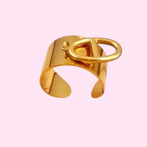May include: A polished gold-tone cuff bracelet featuring a wide, open-ended band design. Attached to the top of the band is a prominent, horizontally oriented oval-shaped charm with a vertical bar running through its center.
