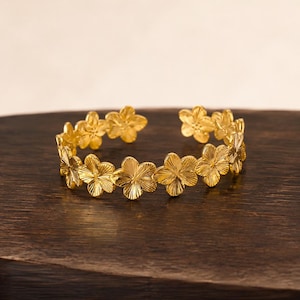 Op de afbeelding: Een goudkleurige manchetarmband bestaande uit een doorlopende rij met elkaar verbonden bloemen met vijf bloemblaadjes. Elke bloem heeft een textuur met een stralend lijnpatroon op het oppervlak. De armband heeft een open ontwerp.