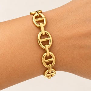 Peut inclure: Un bracelet doré composé d'une série de maillons ovales polis, chacun doté d'une barre horizontale centrale, créant un design de chaîne d'inspiration nautique classique qui entoure le poignet avec une finition métallique lisse et brillante.
