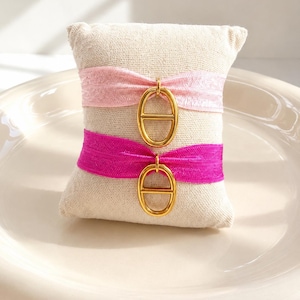 Peut inclure: Un petit coussin beige avec deux élastiques rose et fuchsia. Chaque élastique est orné d'un charm doré de forme ovale. Le coussin est présenté sur une assiette blanche de forme ovale.