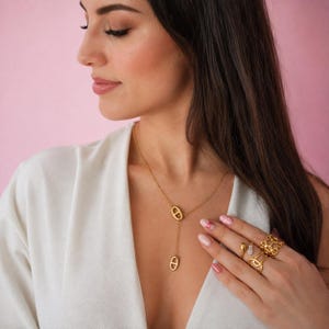 Peut inclure: Un collier lariat doré composé de deux maillons ovales avec une barre horizontale au centre. Le collier est porté avec trois bagues dorées, dont une bague ouverte, une fine bague ornée de petites pierres et une large bague texturée.