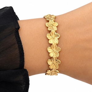 Pode incluir: Uma pulseira em tom dourado com uma série de pingentes de flores de cinco pétalas conectados. Cada flor possui uma superfície texturizada com detalhes radiantes que captam a luz. A pulseira é usada no pulso, exibindo um padrão floral.