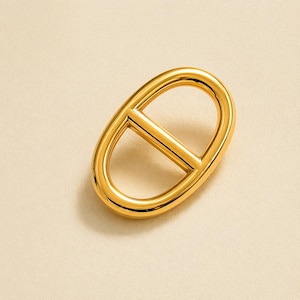 Peut inclure: Boucle de métal doré de forme ovale avec une barre horizontale au centre. La boucle a une finition lisse et polie, sur un fond neutre.