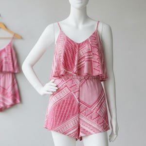 Op de afbeelding: Een roze romper met een geometrisch patroon wordt getoond op een mannequin. De romper heeft een top met V-hals en een gerimpelde overlay en bijpassende shorts. Een andere romper hangt op de achtergrond aan een houten hanger.