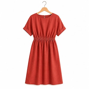 May include: A solid rust-colored dress with short sleeves featuring folded cuffs, a round neckline, and an elasticized waistline that creates a gathered effect. The dress is displayed on a wooden hanger against a plain white background.