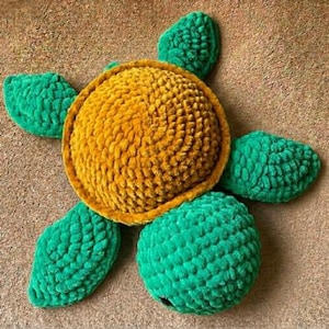 Puede incluir: Un juguete de peluche con forma de tortuga hecho de hilo suave, con un caparazón redondo texturizado de color amarillo dorado y cuatro aletas de color verde vibrante. La cabeza es una forma verde redondeada con un ojo oscuro visible.