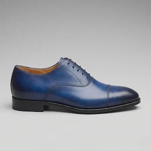 Könnte beinhalten: Ein dunkelblauer Oxford-Schuh aus Leder mit einer Zehenkappe, einem niedrigen Absatz und dünnen schwarzen Schnürsenkeln. Der Schuh hat eine glatte, polierte Oberfläche mit dunkleren Schattierungen an Spitze und Ferse sowie ein beiges Innenfutter.