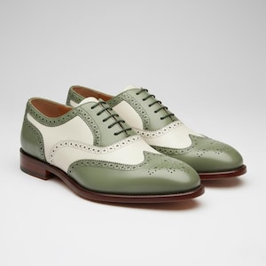 Puede incluir: Un par de zapatos oxford bicolor con paneles de cuero en color verde salvia y crema. Estos zapatos presentan detalles clásicos de brogue con perforaciones decorativas a lo largo de las costuras, cordones verde oscuro y suela de cuero marrón.