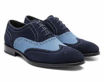 May include: A pair of two-tone oxford shoes featuring a dark navy blue and light blue suede upper with decorative brogue perforations, white contrast stitching, matching navy blue laces, and a solid black sole with a low heel.