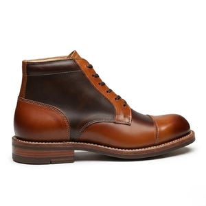 Könnte beinhalten: Zweifarbiges Lederstiefel in Brauntönen, mit einer runden Spitze und Schnürung. Der Stiefel hat einen gestapelten Absatz und eine robuste Sohle, geeignet für verschiedene Anlässe. Das Design des Stiefels ist klassisch.