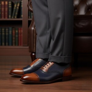 May include: Two-tone leather dress shoes with brown cap toes and heels, navy blue sides, and brown lace panels. The shoes are worn with grey trousers and are set on a wooden floor. The shoes are a classic style.
