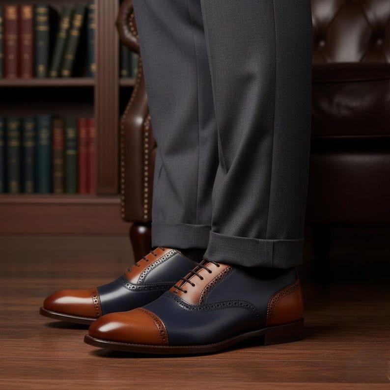 May include: Two-tone leather dress shoes with brown cap toes and heels, navy blue sides, and brown lace panels. The shoes are worn with grey trousers and are set on a wooden floor. The shoes are a classic style.