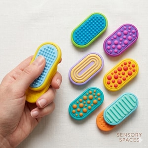 May include: A hand holding a yellow and blue sensory toy, with a textured surface. Six additional colorful sensory toys are arranged on a white surface. The toys are oval-shaped and feature various textures and colors, including blue, purple, orange, and green.